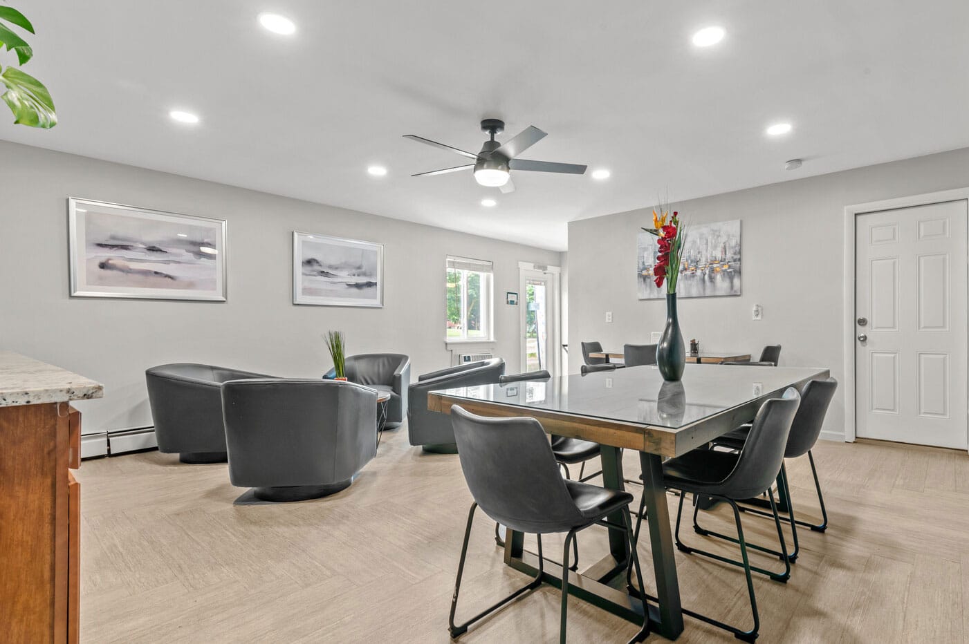 Modern living room with gray chairs, large table, artwork, and ceiling fan.