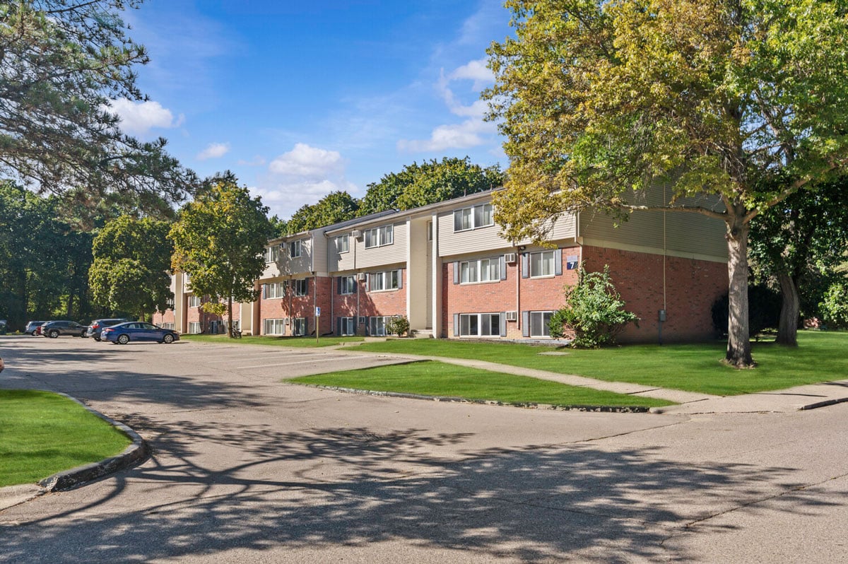 Apartment building with parking lot, surrounded by trees on a sunny day.