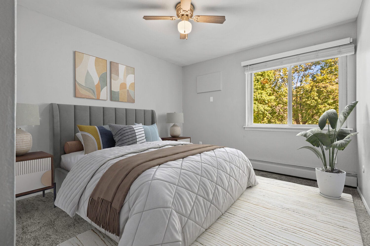Modern bedroom with a large bed, abstract art, and a potted plant by a window with a view of trees.
